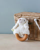 A Senger Naturwelt Sheep Teether, a soft, plush toy shaped like a white lamb with an organic cotton lamb teether made from cherry wood, is placed in front of a woven basket with a white handle. The background is a light blue wall. Handmade in Germany, this charming accessory offers both comfort and function for little ones.