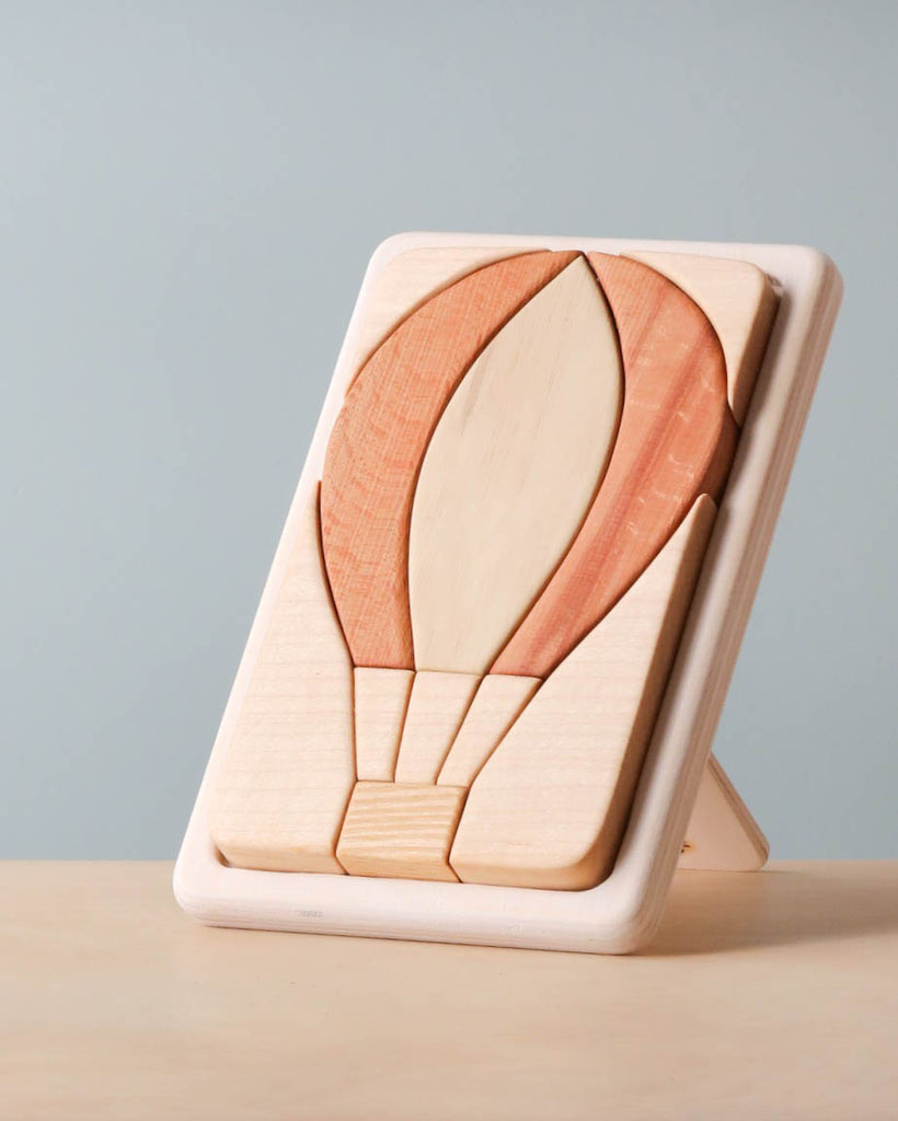 A Handmade Wooden Puzzle depicting a hot air balloon stands upright on a light-colored surface against a plain background. Handmade in Russia, the smoothly crafted puzzle pieces feature two shades of cherry wood, enhanced with linseed oil, giving a minimalist and natural aesthetic.