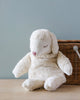 A Senger Naturwelt Cuddly Animal - White Sheep made from organic cotton with a soft white texture sitting in front of a woven basket against a light blue background. The sheep has closed eyes and a peaceful expression.