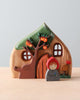 The Handmade Wooden Fairy Cottage boasts a whimsical design, featuring a tree adorned with orange fruits. In front of the cottage stands a small wooden doll donned in a red hooded cloak. The cottage itself is characterized by round windows and a curved door, with hand-painted details including a small pumpkin and grass nearby.