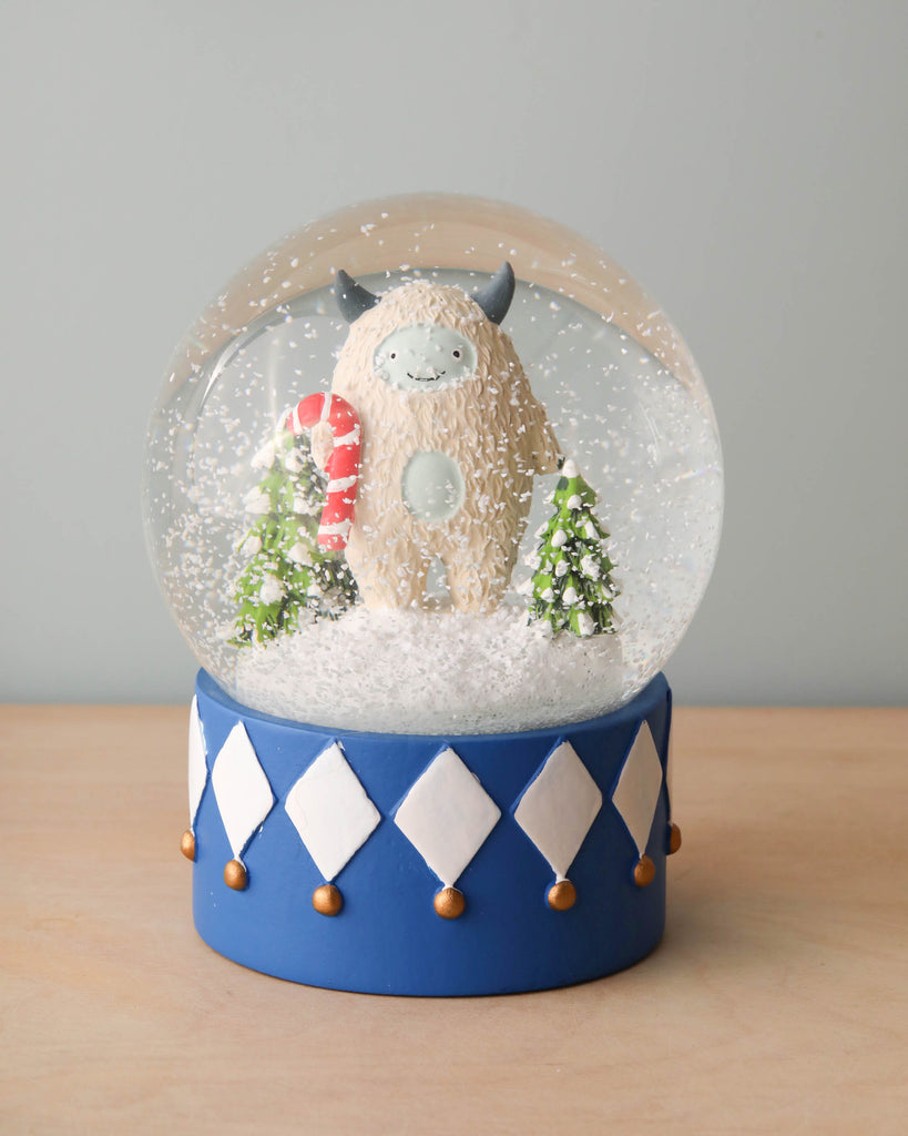 Introducing the Yeti Snow Globe: Enclosed within this charming globe is a cute yeti holding a candy cane, accompanied by two miniature Christmas trees. Delicate snowflakes swirl around the scene. The base is adorned with a blue and white diamond pattern, elegantly set against a soft gray background.