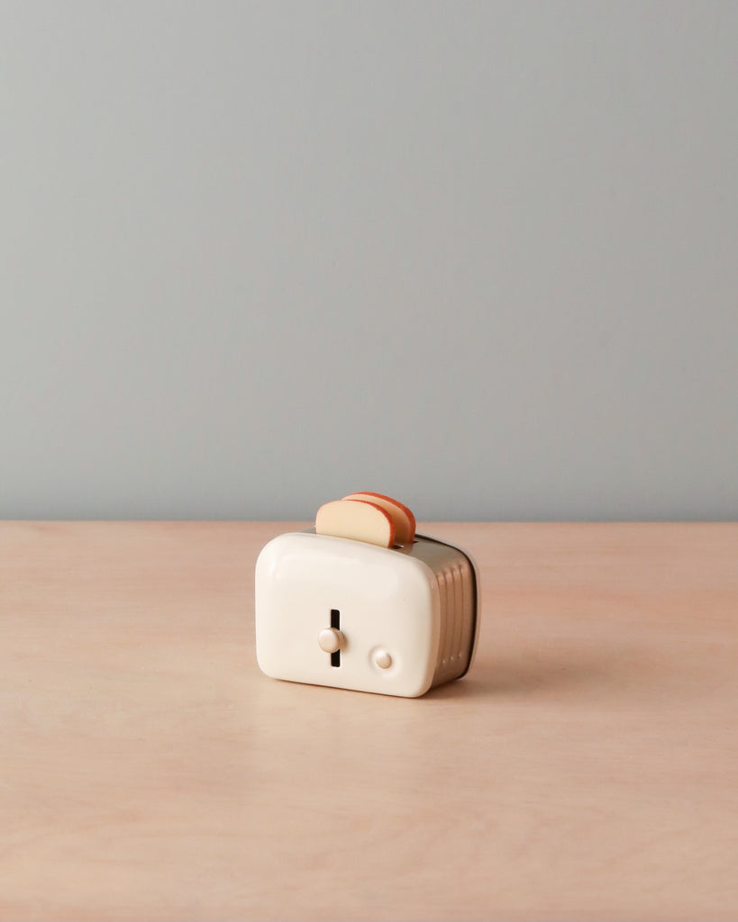 A vintage cream-colored Maileg | Miniature Toaster with a single orange slice of toasted bread on top, sitting on a wooden surface against a plain light blue background.