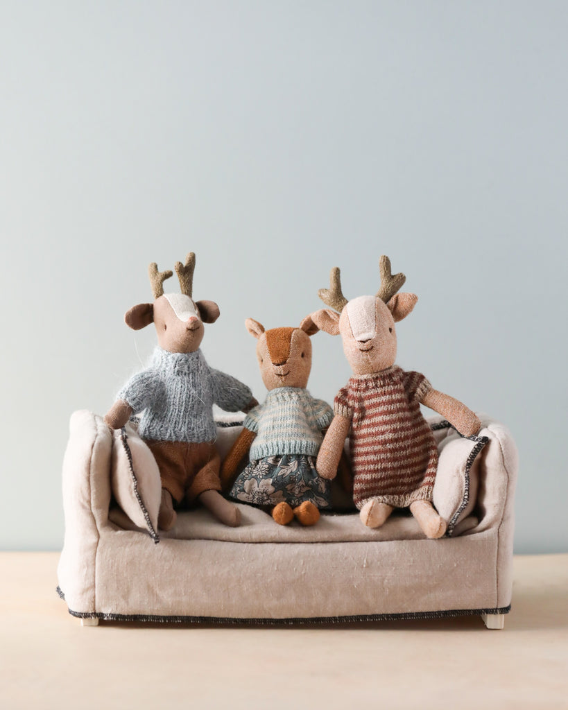 Three small deer stuffed animals each wearing outfits, sitting on a couch together. 