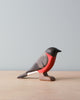 A Handmade Wooden Bullfinch Bird, painted with non-toxic water based paint to resemble a bullfinch, stands on a light wooden surface against a pale blue background.