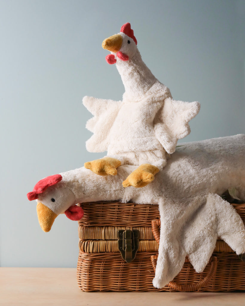 Two Senger Naturwelt Chicken Cuddly Animals, one upright and another lying on its side, atop a wicker basket against a pale blue background. The toys, crafted from organically grown cotton, are white with red.