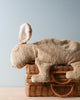A Senger Naturwelt Cuddly Animal - Rabbit made from organic cotton resting on a woven wicker basket with a light blue background. The bunny is in a lying position, showcasing its soft, textured fur and floppy ears.