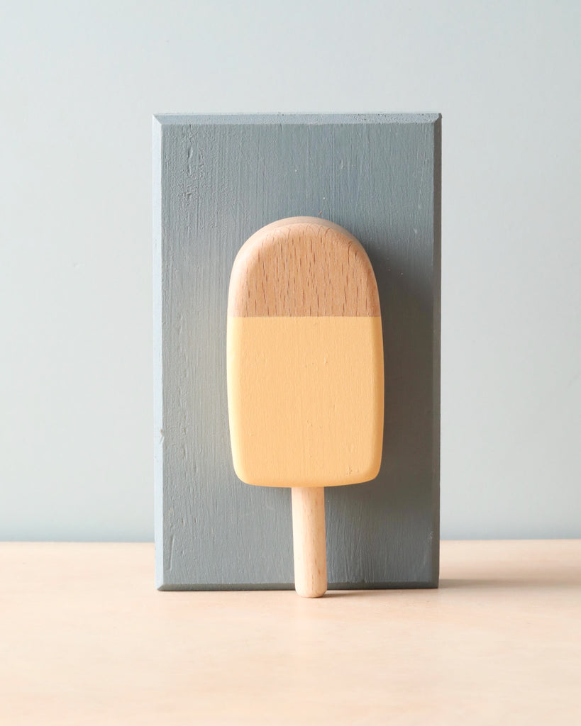 Wooden handmade popsicle in light yellow color (mango flavor). Leaning agains light blue wooden block. 
