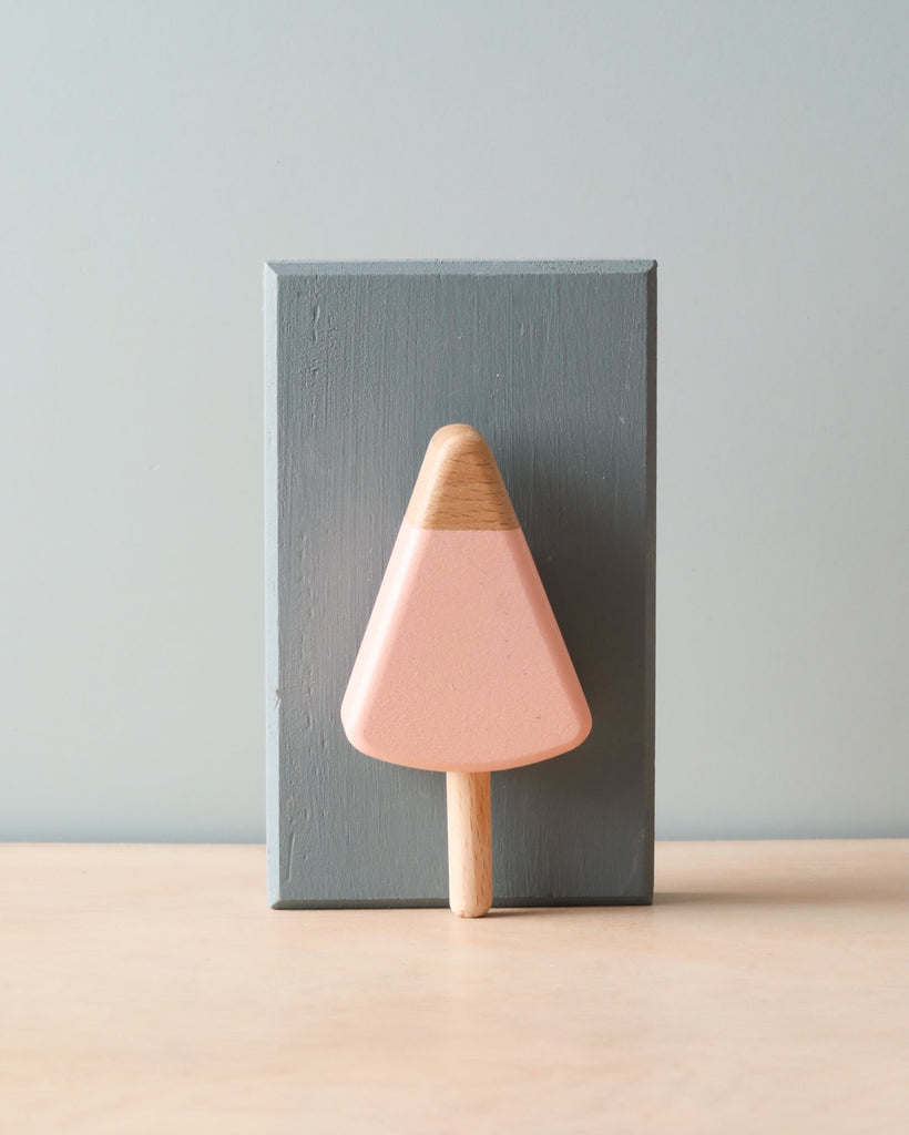 Wooden handmade popsicle in light pink (strawberry flavor). Leaning agains light blue wooden block. 