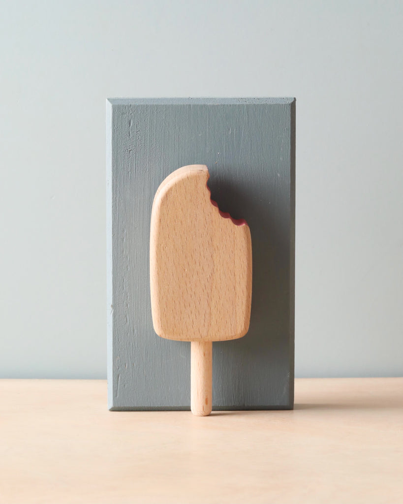 Wooden natural color popsicle (chocolate flavor). Leaning against light blue wooden block. 