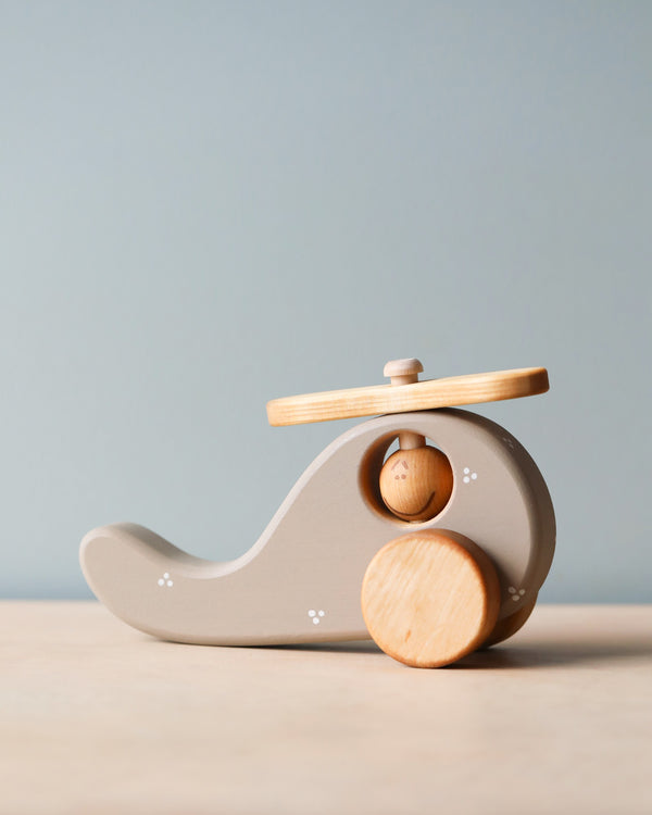 A Handmade Wooden Helicopter with a natural wood finish, made from sustainably harvested birch wood. The helicopter features a rounded body, a propeller on top, and two wheels at the base, all in light brown. It is placed on a plain surface with a light blue background.