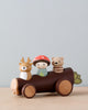 A Timber Taxi, painted brown with leaf details, holds three Merrywood folks: a person in a red hat, and two animals, a deer and a bear, against a plain, light blue