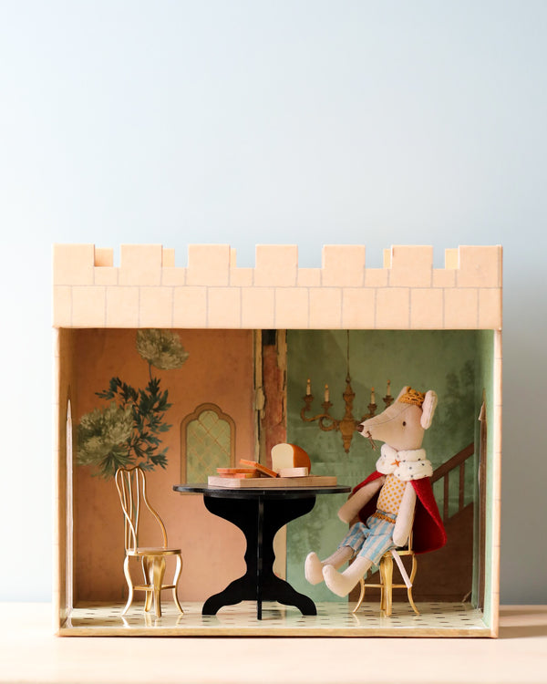 A miniature staged scene with a Maileg Castle Hall mouse sitting at a small table inside a box decorated to resemble a cozy room, complete with painted walls and furniture.