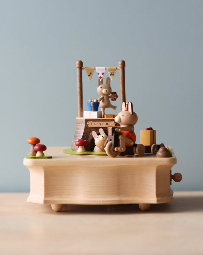 Miniature wooden bunnies on a hand-cranked music box, enjoying a party scene with a "happy hour" banner, drinks, and cakes, against a soft blue background.