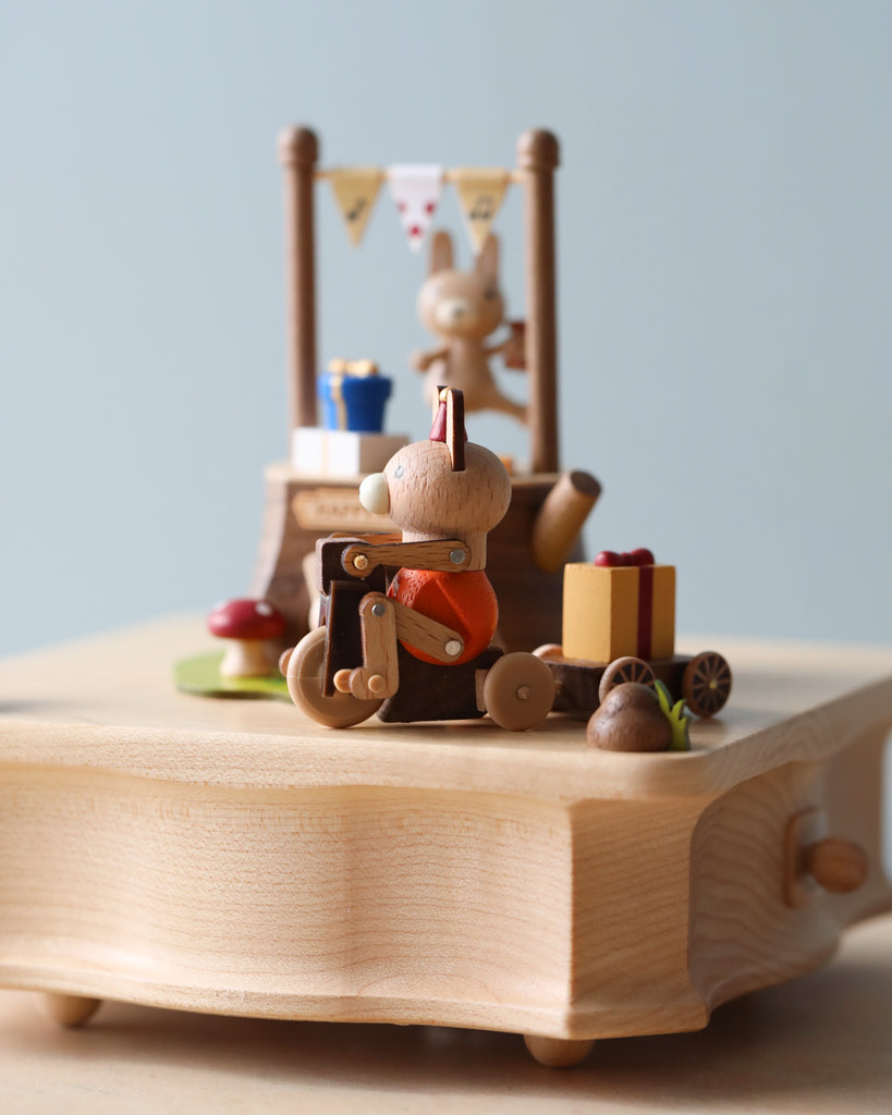 A Wooden Bunny Music Box, crafted from sustainably sourced wood, featuring a small figure in orange taking a photo, a cake with a number one on top, and colorful decorations on a light wooden table.