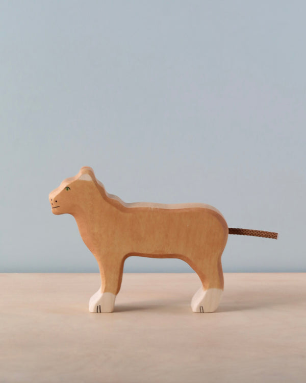 A handcrafted Holztiger Lioness wooden figurine, featuring white paws and a brown string tail, stands on a light surface with a pale blue background, highlighting its quality European craftsmanship.