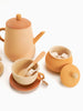 A minimalist Handmade Wooden Tea Set - Flower featuring a peach-colored teapot, cup, sugar bowl, and creamer with a wooden spoon, all on a clean, white background.