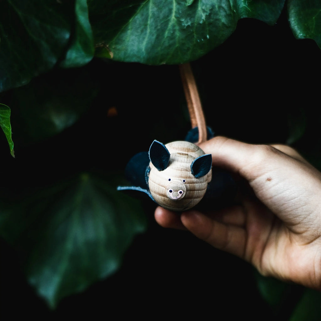 A hand holding a small, hand-painted Handmade Wooden Bat ornament against a dark, leafy green background.