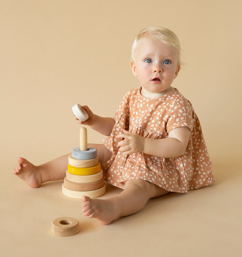 A toddler with blonde hair and blue eyes sits on a beige background, wearing a dotted dress and playing with a Raduga Grez Handmade Pyramid Tower Stacker in Sand.