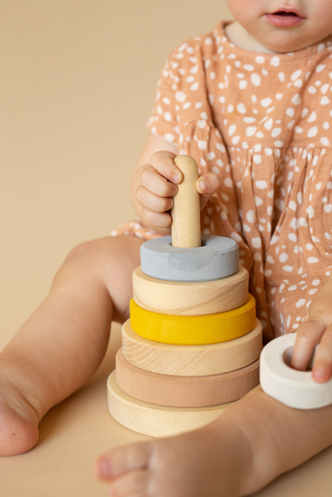 A toddler in a dotted dress plays with a Raduga Grez Handmade Pyramid Tower Stacker - Sand, focusing on placing the rings on a central pole. Only the child's hands and the toy are visible.