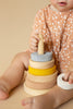A toddler in a dotted dress plays with a Raduga Grez Handmade Pyramid Tower Stacker - Sand, focusing on placing the rings on a central pole. Only the child's hands and the toy are visible.