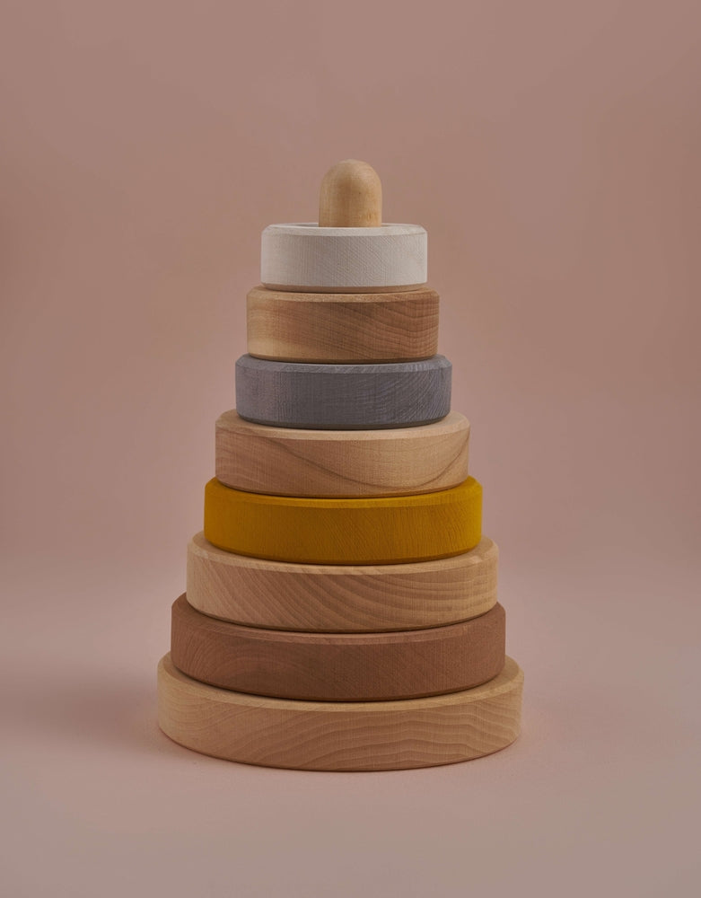 A Raduga Grez Handmade Pyramid Tower Stacker - Sand consisting of a central pole and colorful rings in shades of beige, yellow, gray, and brown—hand-painted with non-toxic paint—against a soft pink background.