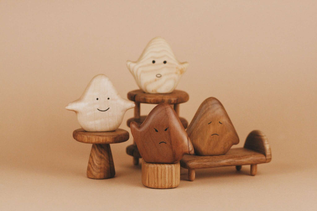 A collection of Wooden Ghost Emotions, each displaying basic emotions with a unique, simple expression, is arranged on various wooden stands and chairs against a beige background. The figures are made from different shades of wood and finished with natural linseed oil, giving them a handcrafted appearance.