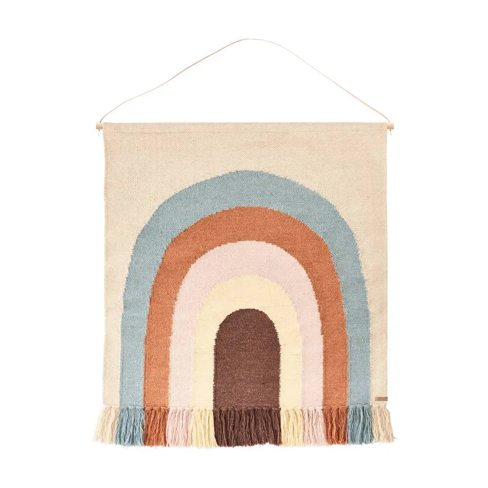 A Follow The Rainbow Wall Rug featuring a multicolored abstract rainbow rug design with fringed ends, suspended from a wooden rod. The colors include brown, blue, pink, and cream.