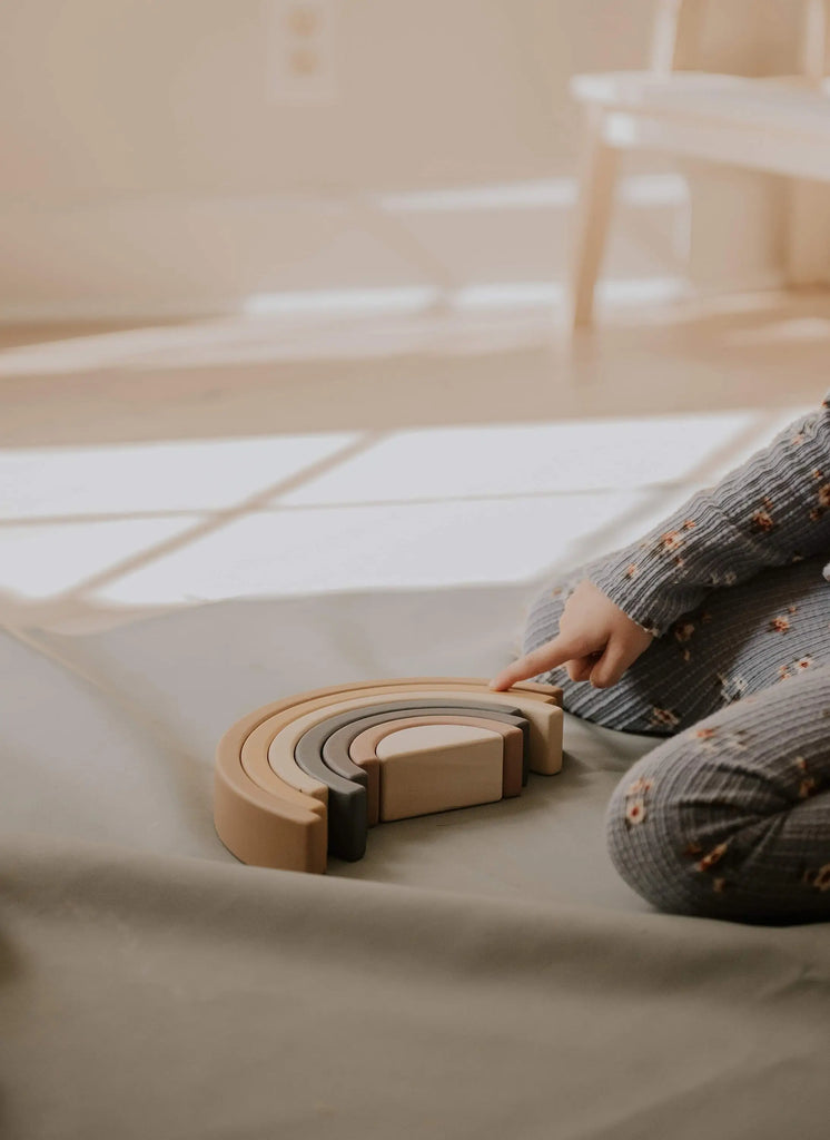 A child wearing patterned pajamas sits on the floor and plays with a Handmade Rainbow Stacker - Mustard. Warm, natural light filters in through a nearby window, creating a cozy atmosphere.