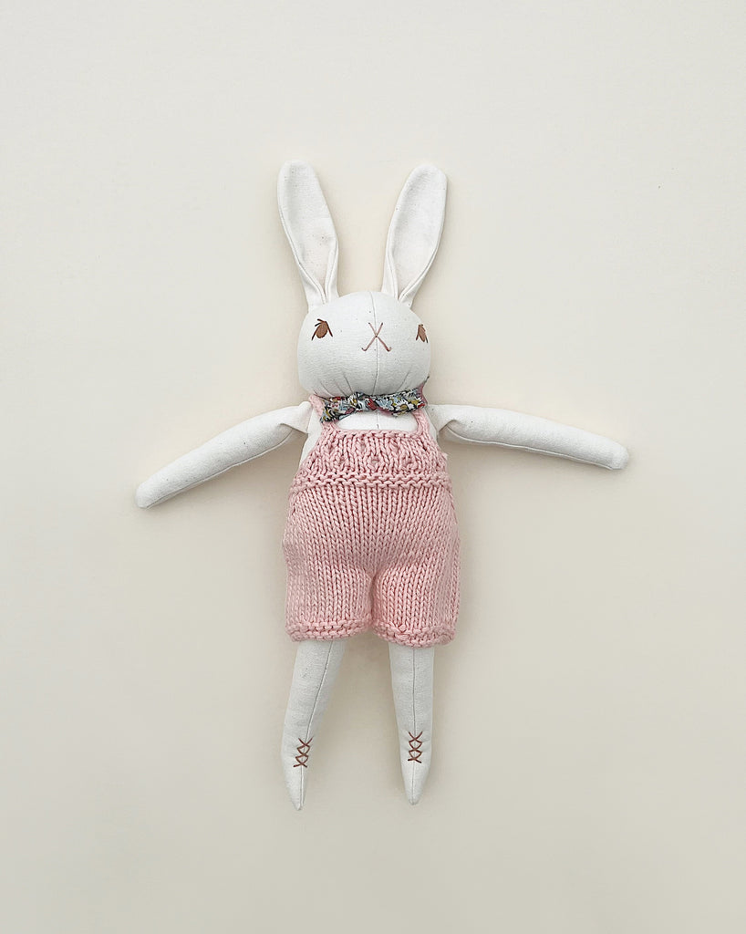 A Polka Dot Club Cream Rabbit in Hand Knit Overalls toy with white and pink features, lying flat against a neutral background.