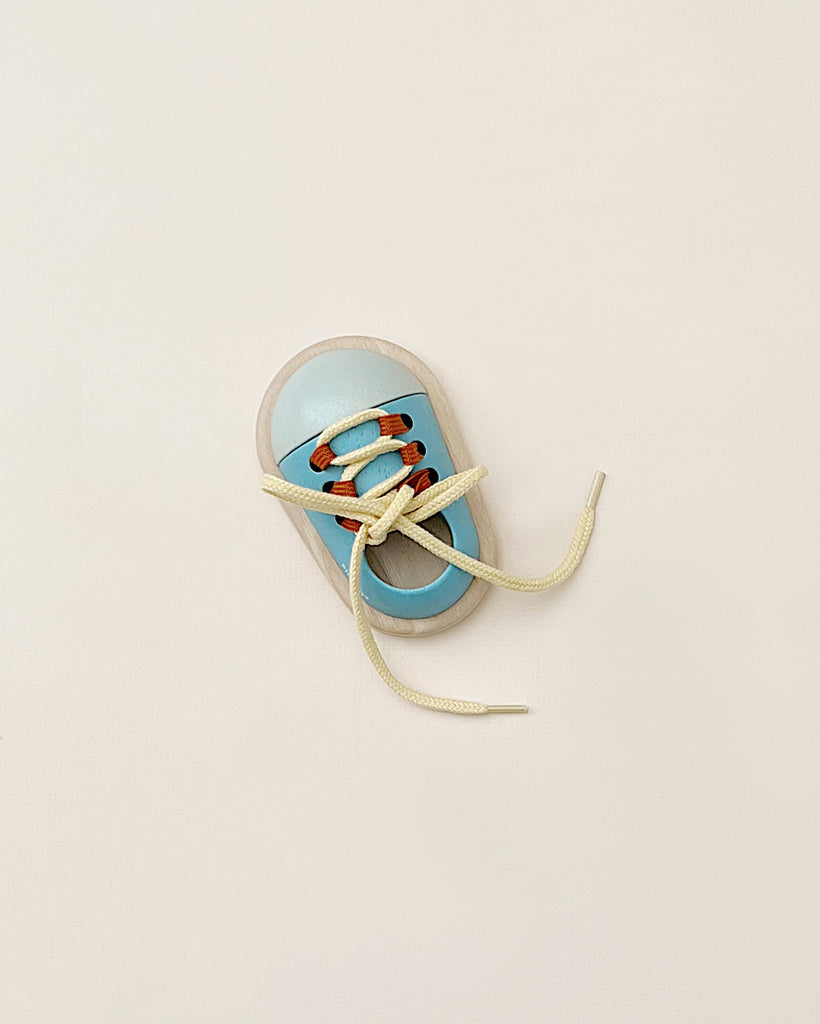 The Tie Up Shoe, a toy toddler shoe in blue and beige with orange accents, is featured against a simple light backdrop. Crafted to enhance hand-eye coordination, it includes an untied beige lace that encourages children to practice tying shoes.