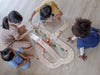 Four young children, diverse in appearance, sit on a wooden floor playing with the Wooden Road System Deluxe, focusing intently on assembling the tracks together.