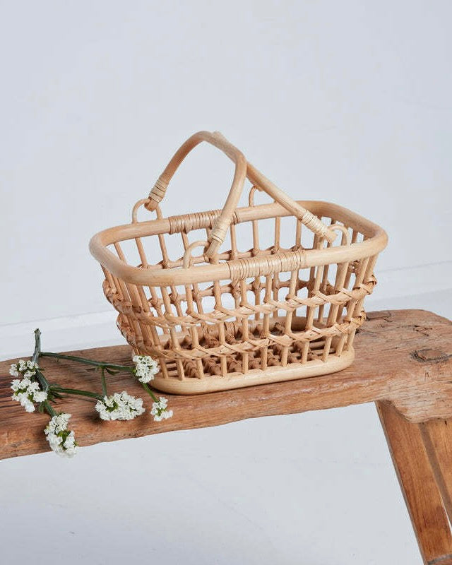 Natural color pretend play shopping basket sitting on wooden bench and white flowers laying next to it. Light gray background. 