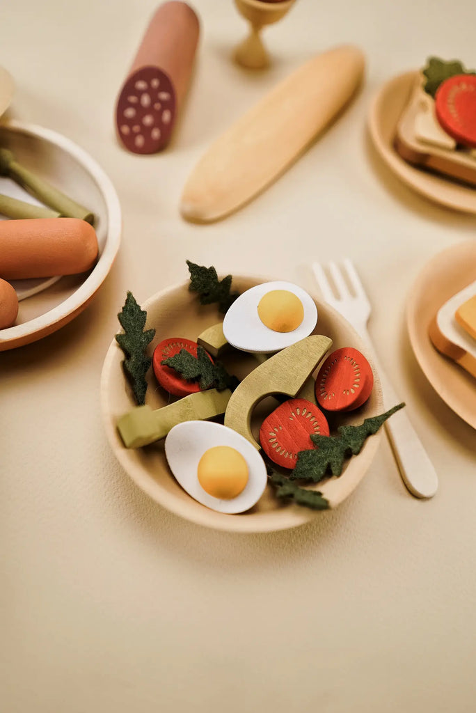 A playful arrangement of Sabo Concept Handmade Wooden Breakfast Set on small plates, featuring items like eggs, tomatoes, and leafy greens painted with non-toxic water-based paint, set against a cream-colored background.