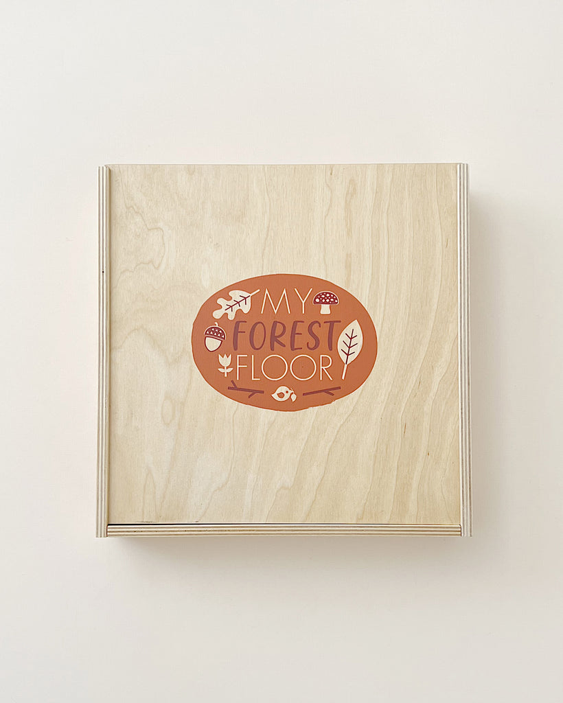 A light wooden square board titled My Forest Floor designed for open-ended play, featuring illustrations of mushrooms, leaves, and wildlife silhouettes within an oval inset.
