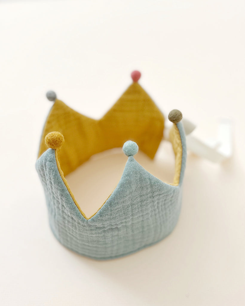 A soft organic cotton Reversible Crown with alternating sections of mustard yellow and teal blue, adorned with colorful pom-poms on the tips, displayed against a light background.