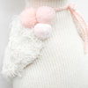 Close-up of a Cuddle + Kind Harlow The Swan with pink fluffy pom-poms attached by a pink string, hand knit to highlight a textured and cozy winter fashion detail.