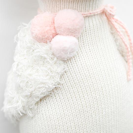 Close-up of a Cuddle + Kind Harlow The Swan with pink fluffy pom-poms attached by a pink string, hand knit to highlight a textured and cozy winter fashion detail.