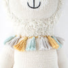 Close-up of a Cuddle + Kind Llama Stuffed Animal with a smiling face, featuring a fluffy white scarf adorned with colorful tassels in shades of gold, teal, and gray.