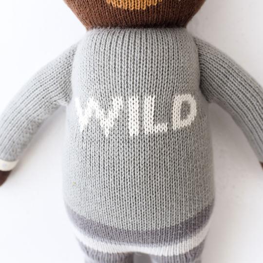 A close-up image of a Cuddle + Kind Oliver the Bear toy with a grey sweater that has the word "wild" in white letters on its back. The texture of the yarn is clearly visible.