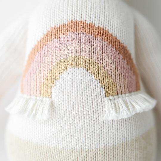 A close-up of a textured, white hand-knit fabric with a decorative rainbow design in pastel pink, beige, and peach colors, accented by small white fringes at the ends, featuring Cuddle + Kind Zara the Unicorn.