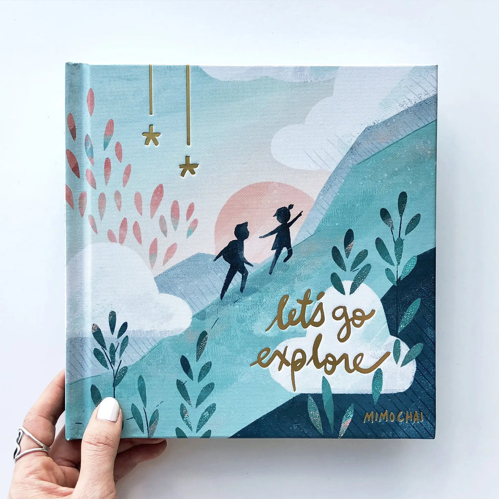 A hand holding a hardcover illustrated Let's Go Explore Book with an illustration of two children playing under a stylized sunset, surrounded by plants and hanging stars.