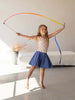 A young girl in a polka dot top and blue skirt joyfully engages in pretend play with Sarah's Silk Rainbow Streamer Wand in a brightly lit room, standing barefoot with a smile.