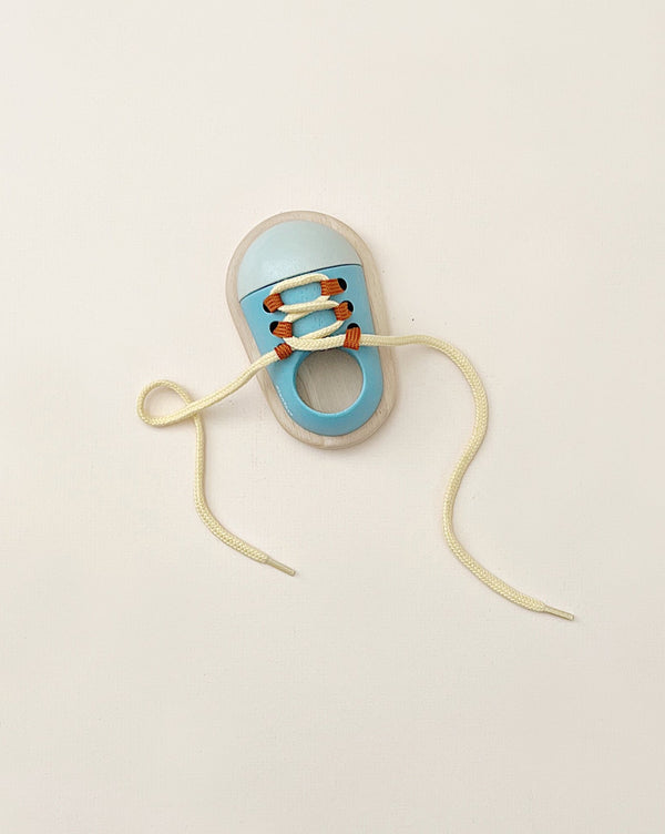 The Tie Up Shoe, featuring a compact design in light blue with white laces and orange accents, is displayed on a beige background. The round opening and partially knotted laces provide an opportunity to enhance fine motor skills as one loose end extends outward.