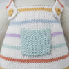 Close-up of a Cuddle + Kind Lamb Stuffed Animal with a pastel striped pattern and a pale blue pocket on the front. The texture of the yarn is prominently visible.