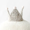 A close-up image of a Cuddle + Kind Harlow The Swan hand-knit hat with three small pompoms on top, placed on a white rounded surface, providing a minimalist aesthetic.