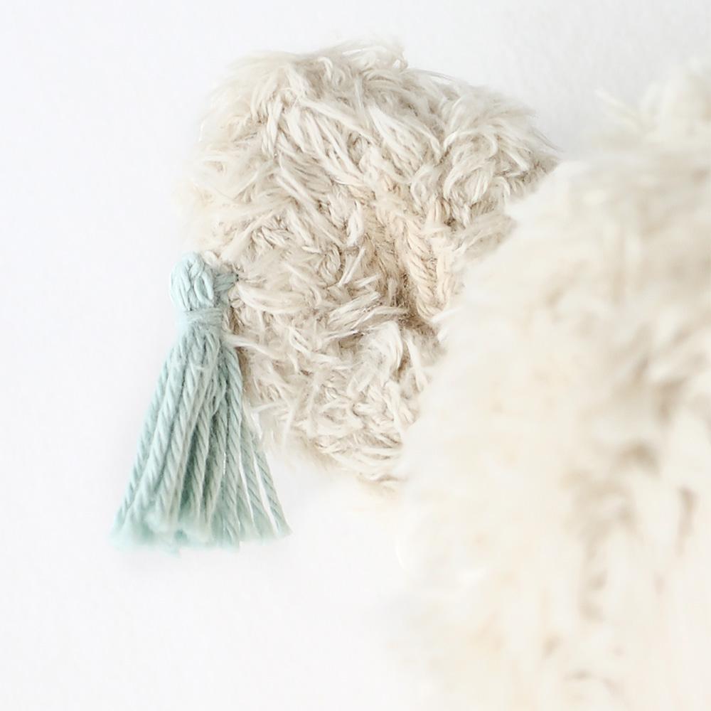 A close-up of a hand knit Cuddle + Kind Llama Stuffed Animal against a textured white furry background, showcasing details of the yarn and the softness of the materials.