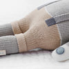 Close-up of a Cuddle + Kind Dog Stuffed Animal made of patchwork fabric in beige and grey hues, lying on a soft white surface. The toy features stitched details and is filled with hypoallergenic polyfill.