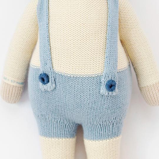 A close-up image of a Cuddle + Kind Lamb Stuffed Animal wearing blue overalls and featuring detailed stitching and two buttoned shoulder straps. The background is plain and light-colored, emphasizing the doll.