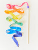 A Sarah's Silk Rainbow Streamer Wand with pink to blue streamers cascading beside a white stick, topped with a purple ball, against a white background.