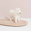 A small, fluffy Hand Felted White Wool Sheep figurine, handcrafted from naturally-dyed wool with a simple, smiling face stands on a light wooden oval base against a soft pink background.